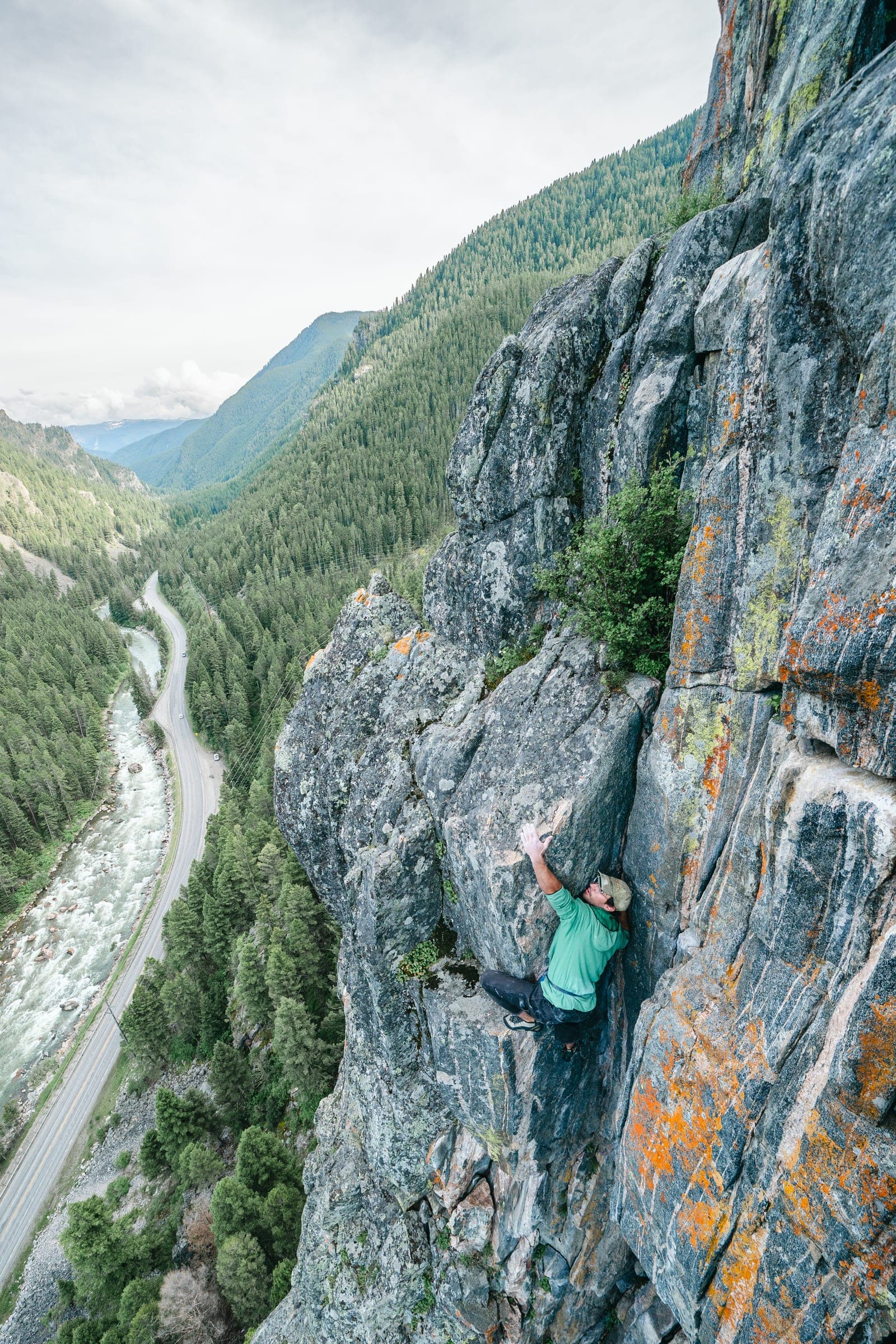 Climber on an orange-lichen arete high above a forested valley and road