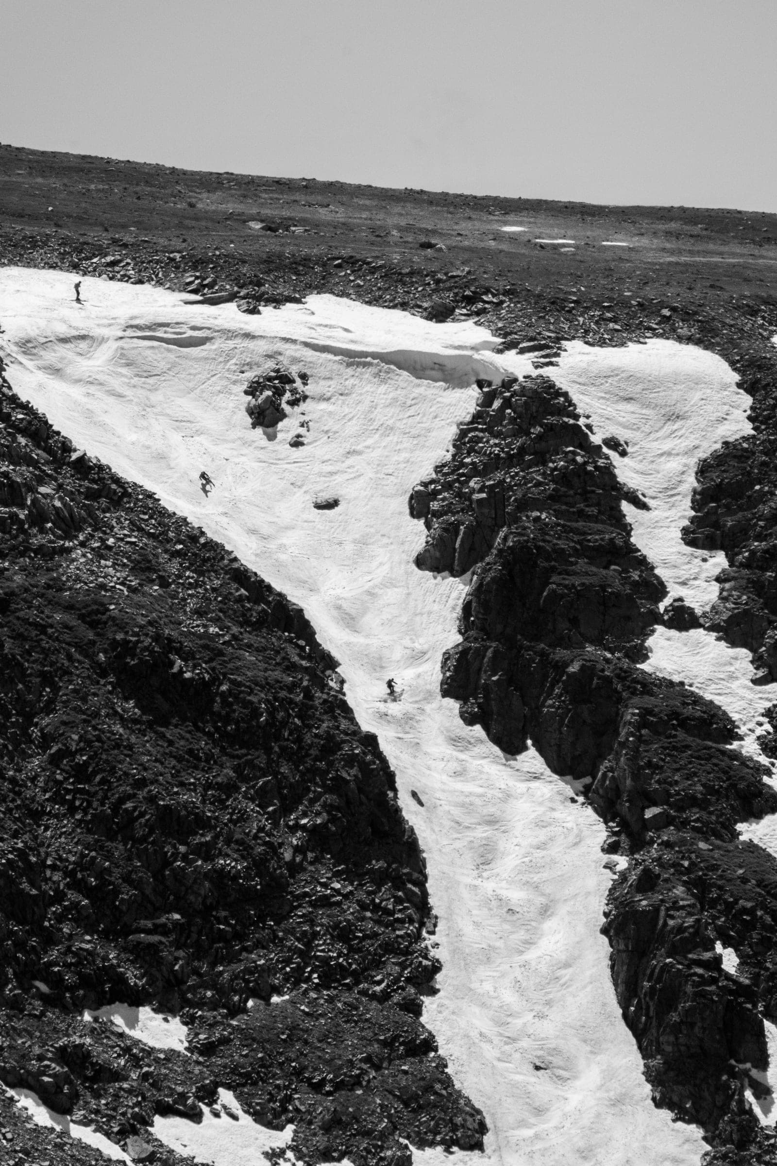 B&W couloir with tiny skiers carving through dark rock bands
