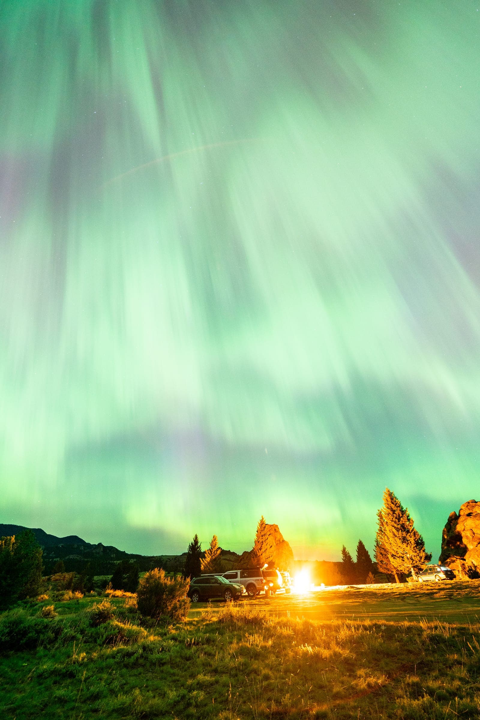 Green aurora curtains over glowing campfire and backlit trees