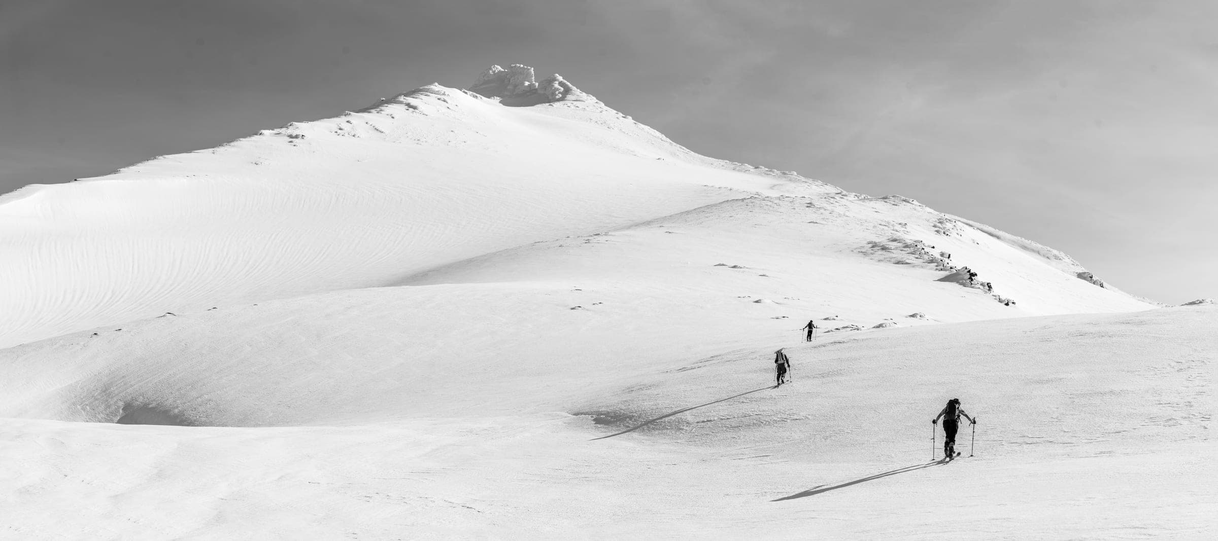 B&W panorama: three tiny skiers on a vast snow dome