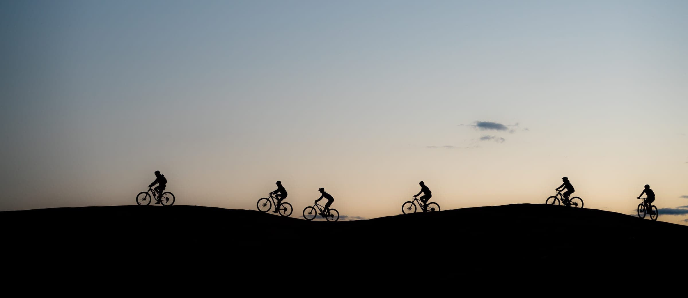 Six mountain bikers silhouetted along a desert ridge at dusk