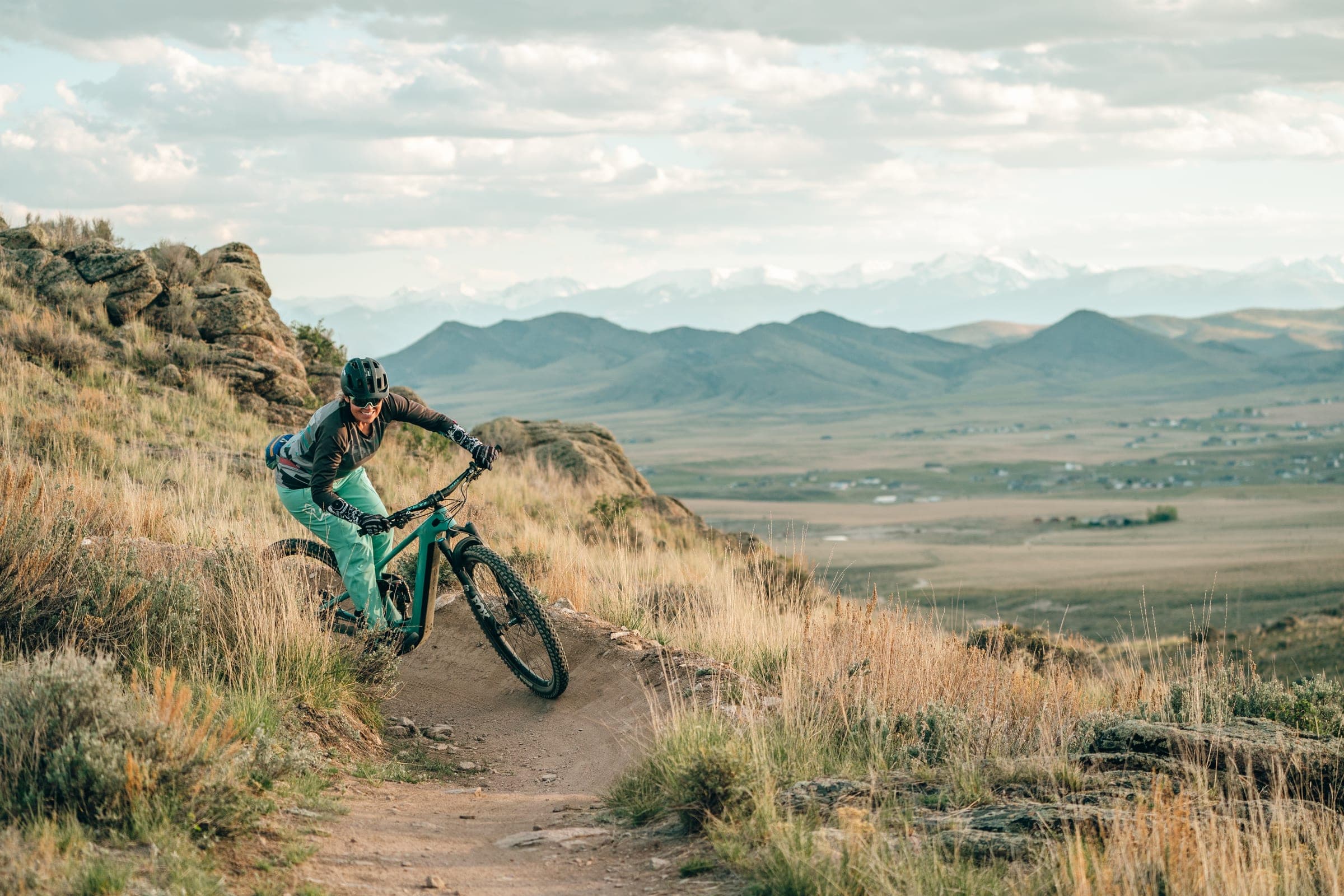 Rider in turquoise pants carving the trail with the valley and range behind