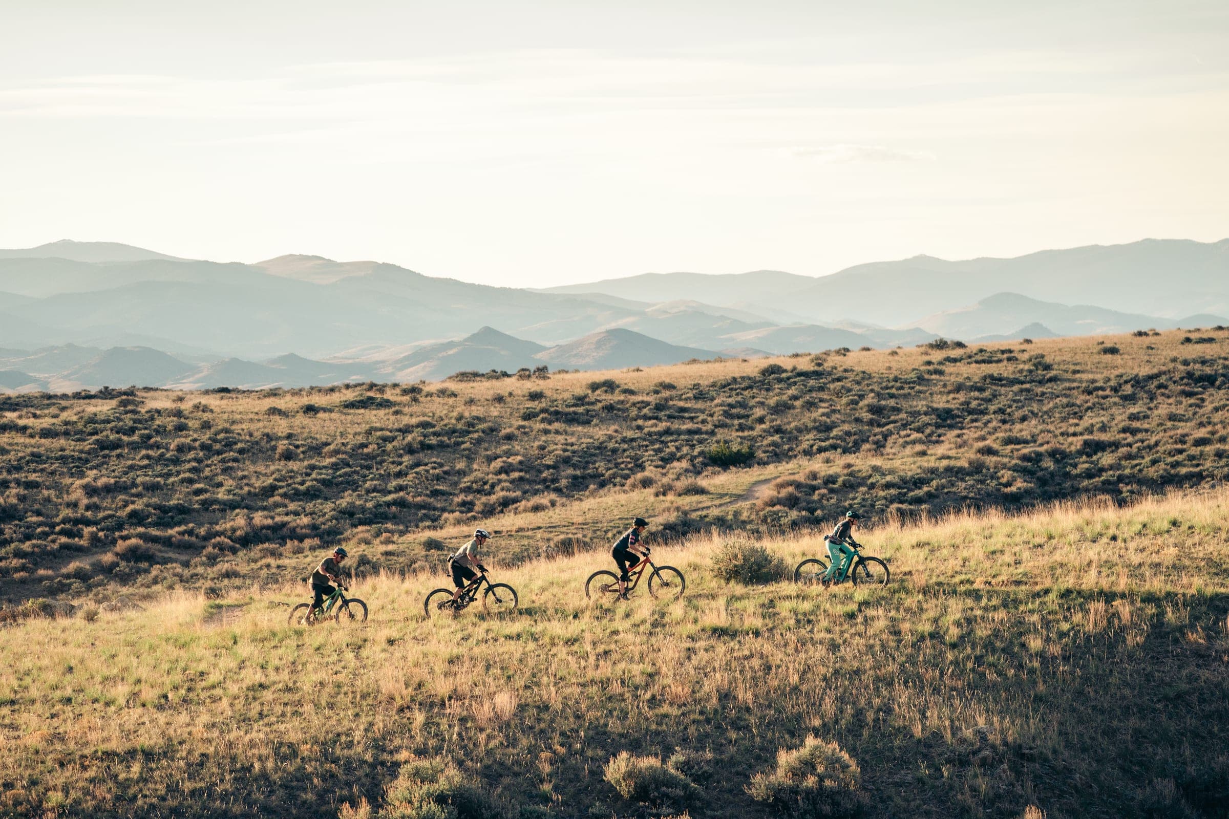 Four mountain bikers crossing a golden sagebrush hillside below hazy layered ranges