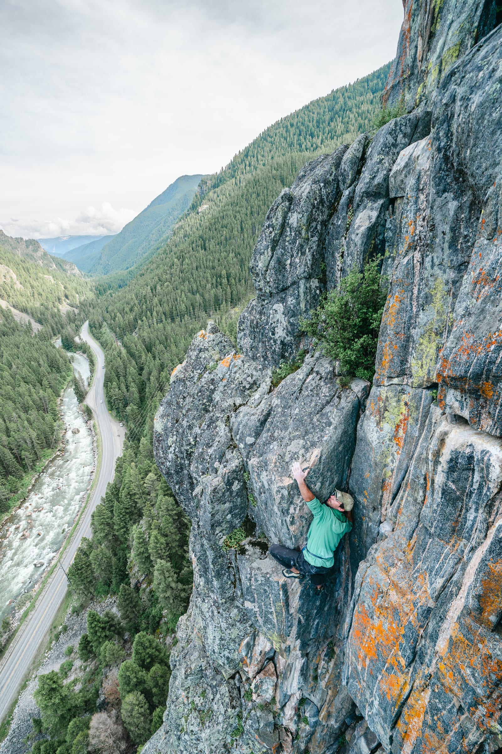 Climber on an orange-lichen arete high above a forested valley and road