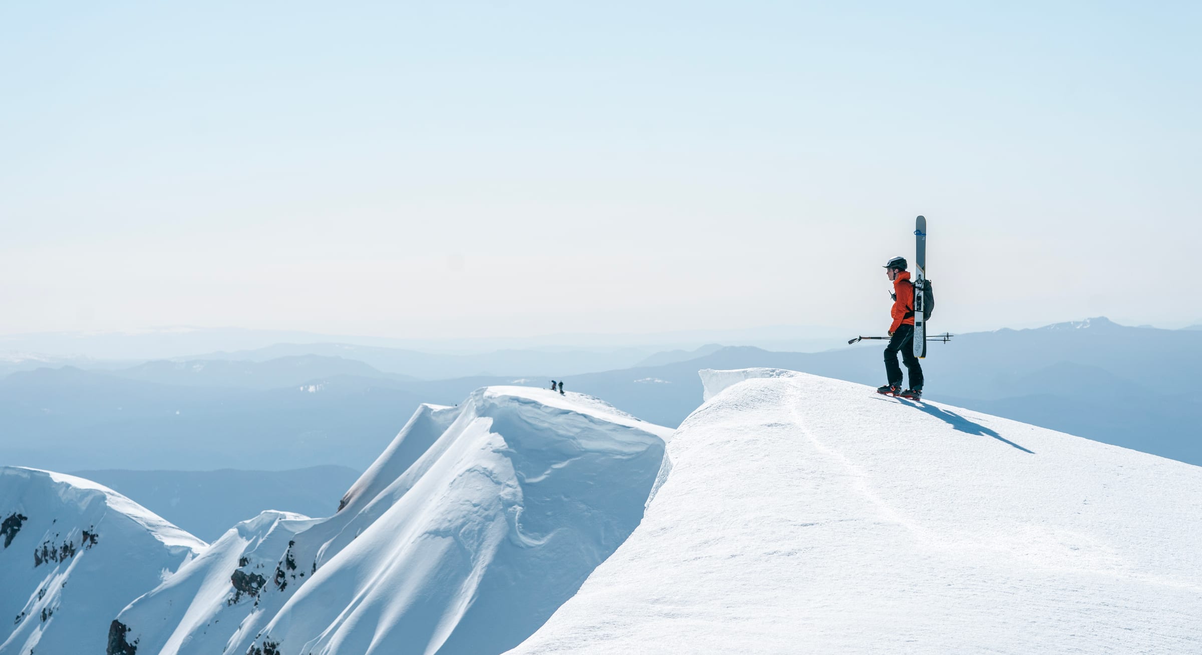 Skier on a cornice summit above a sea of hazy ridges