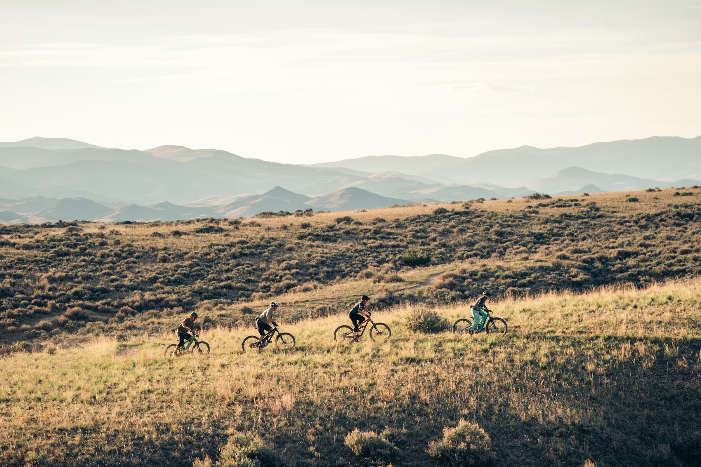 Four mountain bikers crossing a golden sagebrush hillside below hazy layered ranges